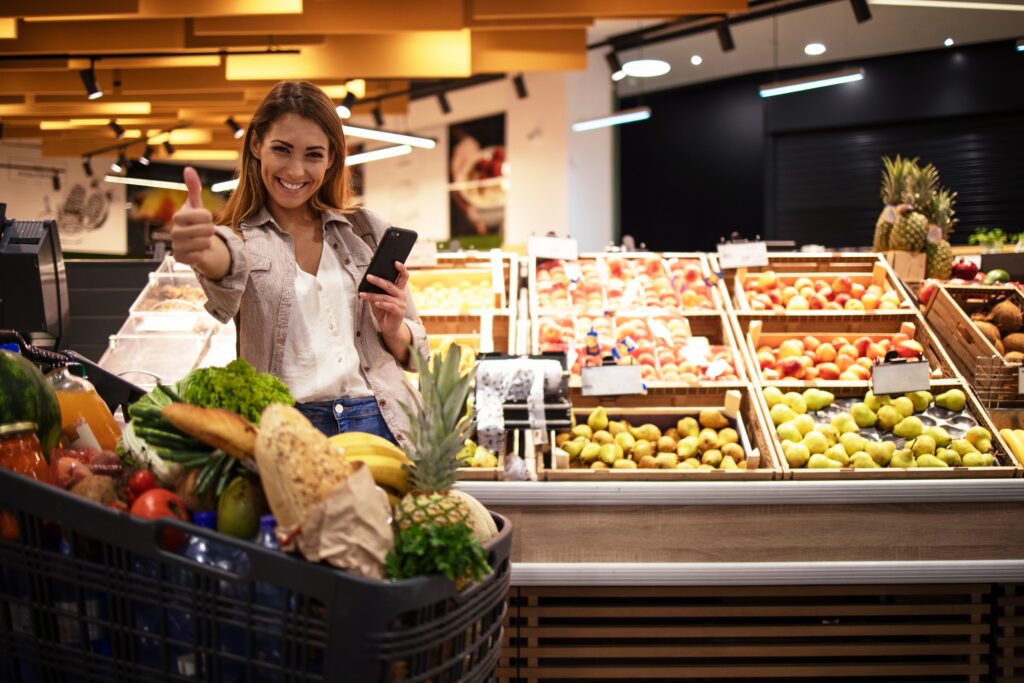 woman-with-smart-phone-in-supermarket-standing-by-the-shelves-full-of-fruit-at-grocery-store-holding-thumbs-up